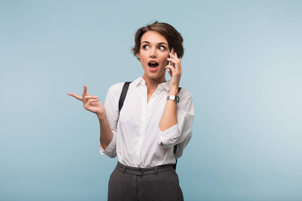 Mujer joven con el pelo corto oscuro en camisa blanca asombradamente mirando a un lado hablando en el teléfono celular sobre fondo azul
 - Foto, imagen