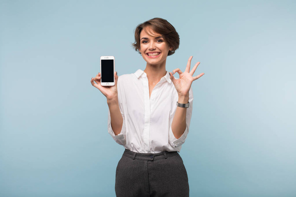 Bardzo radosna dziewczyna z ciemne, krótkie włosy w białej koszuli, trzymając telefon w ręce szczęśliwie Wyświetlono ok gest na aparat na niebieskim tle - Zdjęcie, obraz