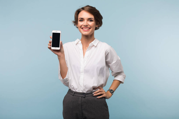 Piękna dziewczyna uśmiechający się ciemne, krótkie włosy w białej koszuli radośnie patrząc w aparacie z telefon w ręce nad niebieskim tle - Zdjęcie, obraz