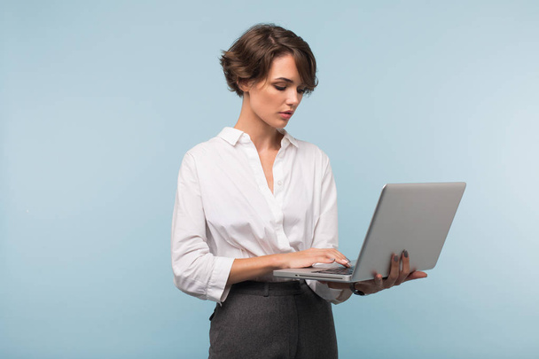 Fiatal, vonzó üzletasszony elgondolkodva dolgozik laptop mint kék háttér fehér ing-sötét rövid haj - Fotó, kép