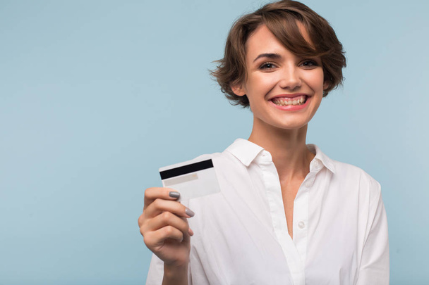 Beautiful smiling girl with dark short hair in white shirt holding credit card in hand joyfully looking in camera over blue background - Photo, Image