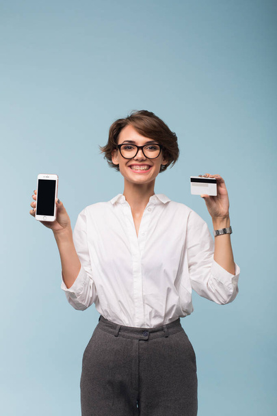 Young smiling woman with dark short hair in white shirt and eyeglasses holding credit card and cellphone in hands happily looking in camera over blue background - Foto, imagen