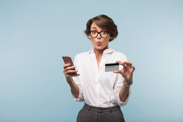 Young pretty woman with dark short hair in white shirt and eyeglasses holding credit card and cellphone in hands amazedly looking in camera over blue background - Φωτογραφία, εικόνα