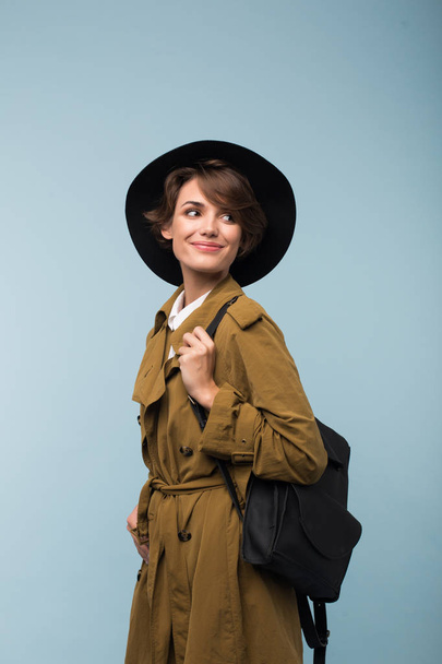 Beautiful smiling girl with dark short hair in trench coat and hat with backpack dreamily looking aside over blue background - Foto, Imagem