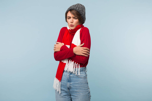 Young woman with dark short hair in red sweater,scarf and knitted hat shaking from cold over blue background - Foto, immagini