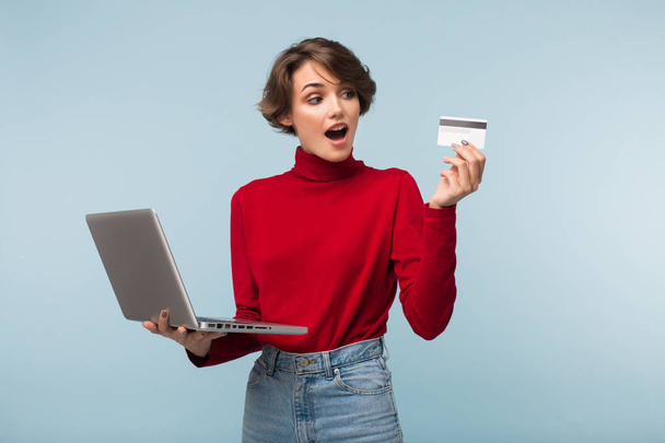 Mujer joven con el pelo corto oscuro en suéter rojo y jeans que sostiene el ordenador portátil en la mano asombradamente mirando en la tarjeta de crédito sobre fondo azul
 - Foto, imagen