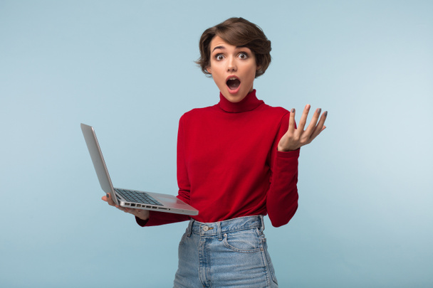 Young shocked woman with dark short hair in red sweater and jeans holding laptop in hand amazedly looking in camera over blue background - Photo, image