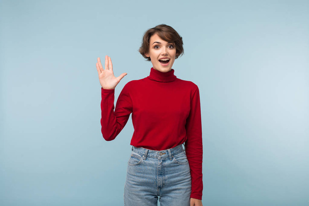 Beautiful cheerful girl with dark short hair in red sweater and jeans showing  vulcan greeting gesture happily looking in camera over blue background  - Fotoğraf, Görsel