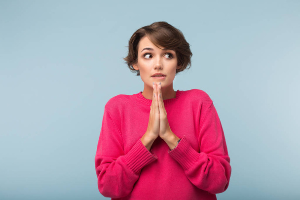 Jovem mulher com medo com cabelo curto escuro em suéter rosa mantendo as mãos em gesto de oração surpreendentemente olhando de lado sobre fundo azul
 - Foto, Imagem