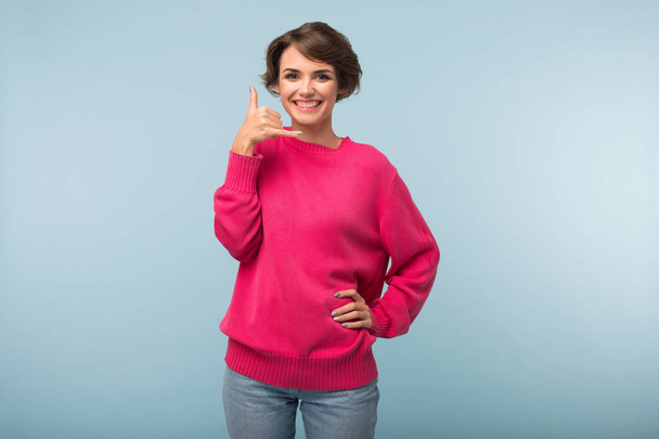 Jovem mulher atraente com cabelo curto escuro em suéter rosa e jeans mostrando gesto telefone alegremente olhando na câmera sobre fundo azul
 - Foto, Imagem