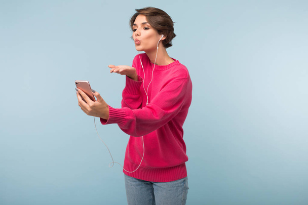 Fiatal, vonzó nő rózsaszín pulóver és a fülhallgató álmodozva küldő légi csók gazdaság cellphone kéz alatt kék háttér - Fotó, kép