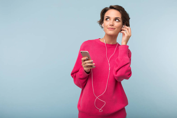 Mujer atractiva joven en suéter rosa sosteniendo el teléfono celular en la mano soñando mirando a un lado escuchando música en auriculares sobre fondo azul
 - Foto, Imagen