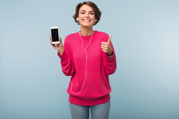 Jeune jolie femme souriante en pull rose et écouteurs tenant le téléphone portable dans la main montrant joyeusement grand pouce geste vers le haut en regardant dans la caméra sur fond bleu
 - Photo, image