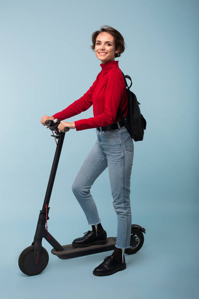 Young pretty smiling girl in red sweater and jeans with black backpack standing with electric scooter happily looking in camera over blue background - Foto, Bild