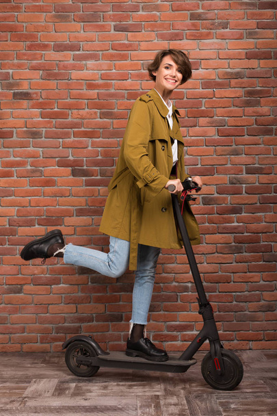 Young beautiful woman in trench coat and jeans standing on electric scooter happily looking in camera over brick wall background - Fotó, kép