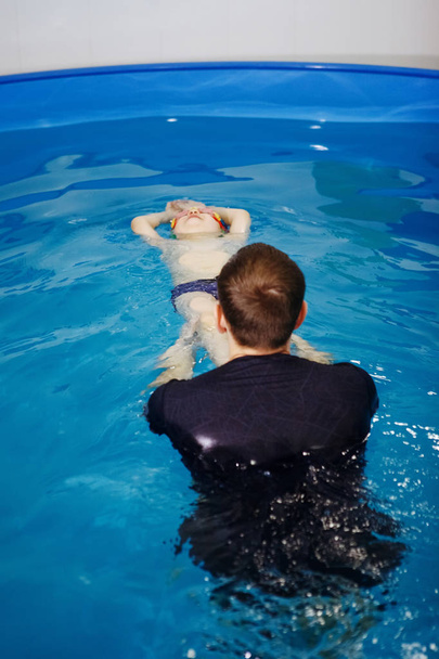 Τρεις-χρονών παιδί με μπλε πώμα και κολύμπι γυαλιά είναι λουσμένο σε μια πισίνα με τη βοήθεια ενός εκπαιδευτή αρσενικό. Aquatherapy. Αποκατάσταση του νερού. Καταδύσεις. Υγιεινός τρόπος ζωής. - Φωτογραφία, εικόνα