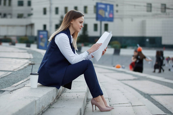 Mulher muito alegre com uma pilha de papel em um terno azul escuro e camisa branca está sentado nos degraus de pedra na cidade de verão
. - Foto, Imagem