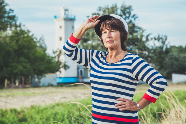 Beautiful elderly woman in a striped dress on the beach at background of old lighthouse - Fotografie, Obrázek