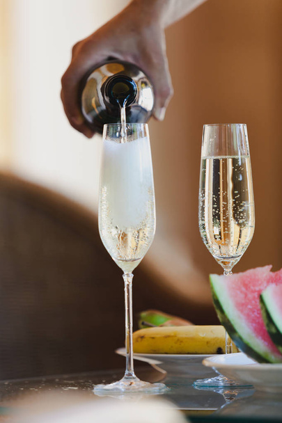 Romantic plot with champagne. The hand pours a sparkling wine with a thin stream into tall, elegant glasses. Next to fruit and watermelo - Photo, image