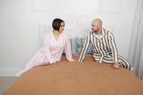Nuori kaunis nainen ja kalju parrakas mies pyjamassa puhuvat sängyssä tyynyillä ja ruskeilla vuodevaatteilla.
. - Valokuva, kuva