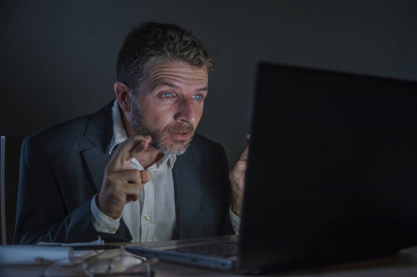 young excited and attractive businessman crossing fingers for luck hoping to win online bet while working late night at office laptop computer desk in internet gambling and money making  - 写真・画像