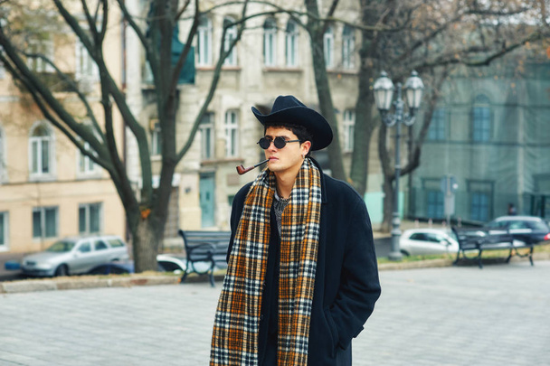 a man smokes a pipe in the city . portrait of a stylish fashionable man on the streets . man in black coat, plaid scarf and hat - Photo, Image
