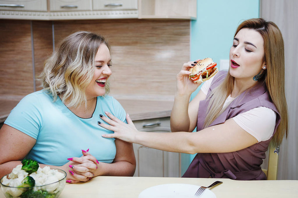 Young pretty fat blonde and her slim girlfriend are taking each other's juicy hamburger and laughing in the kitchen. The concept of healthy eating, diet, fight with overweight. - Foto, Imagen