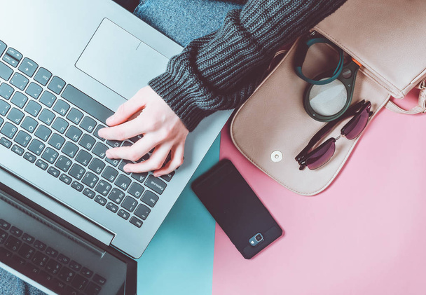 Frauenhände benutzen einen Laptop auf dem Hintergrund einer Tasche, eines Smartphones, eines Armbands und anderer Gadgets und Accessoires. Draufsicht auf pastellfarbenem Hintergrund. - Foto, Bild