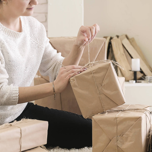 a young brunette woman in a white sweater is sitting on the floor in a bright room and examining boxes of gifts in craft paper.girl opens a box with gifts during the holiday - Фото, изображение