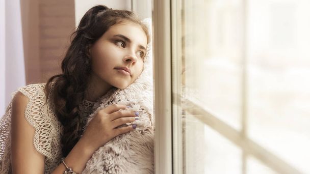 belle fille brune assise sur le rebord de la fenêtre embrassant un oreiller beige doux et regardant par la fenêtre
. - Photo, image