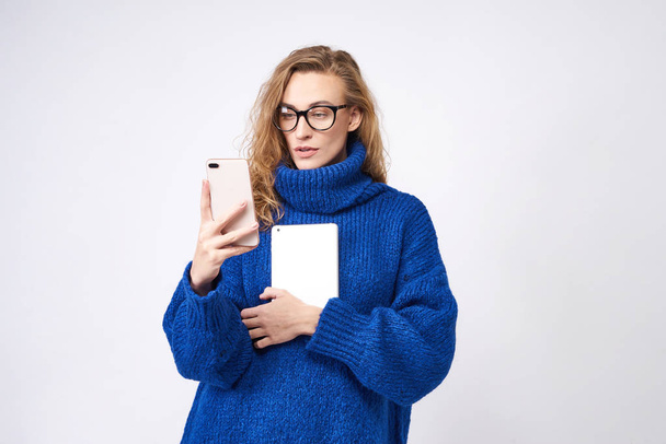 Blonde female student with mobile phone and tablet in hands on white background - Fotó, kép
