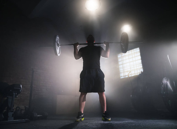 Weightlifter flipping two-handed dumbbell and preparing for workout in gym interior. Cinematic mood with dust, dramatic lightning and smoke. Active and healthy lifestyle. - Fotoğraf, Görsel