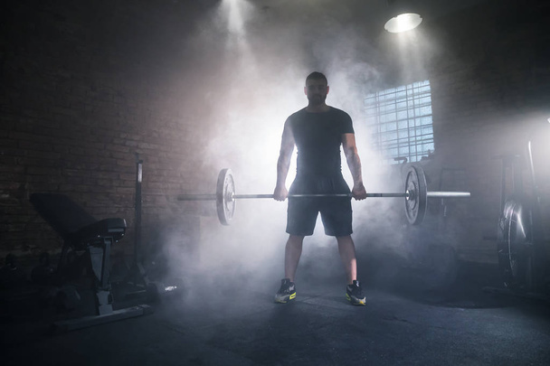 Weightlifter flipping two-handed dumbbell and preparing for workout in gym interior. Cinematic mood with dust, dramatic lightning and smoke. Active and healthy lifestyle. - Fotó, kép