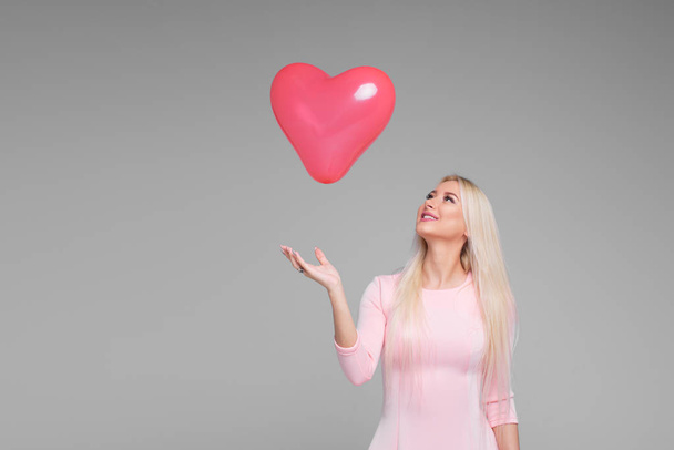 Beautiful young blond woman with pink heart shape air balloon on grey background. Woman on Valentine's Day. Symbol of love - Image. Space for text - Fotoğraf, Görsel