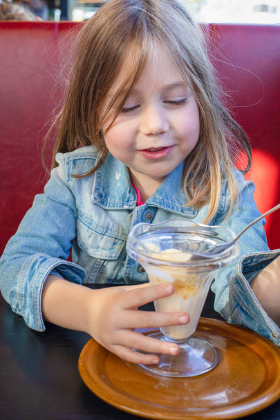 Portrait of five years old blonde little child with blue denim jacket, sitting in red sofa and black table in a restaurant, eating vanilla ice cream with spoon from crystal cup. vertical shot - Фото, зображення