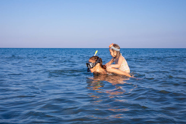 woman with little girl smiling in her back, piggybacking, both with diving glasses to snorkeling, in the sea water of a beach in Andalusia (Spain) - Photo, Image