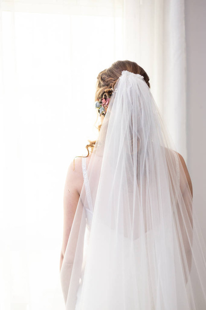 eine Braut auf dem Rücken mit weißem Schleier und Kleid und einer Blume auf ihrem blonden Haar, die aus dem Fenster schaut - Foto, Bild