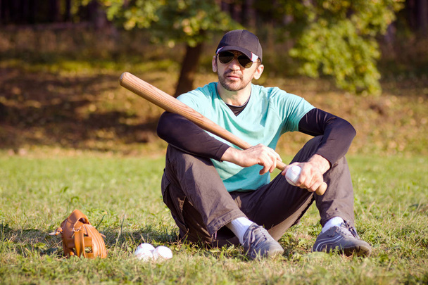 The baseball player is sitting and resting on grass with his sports equipments. - Photo, Image