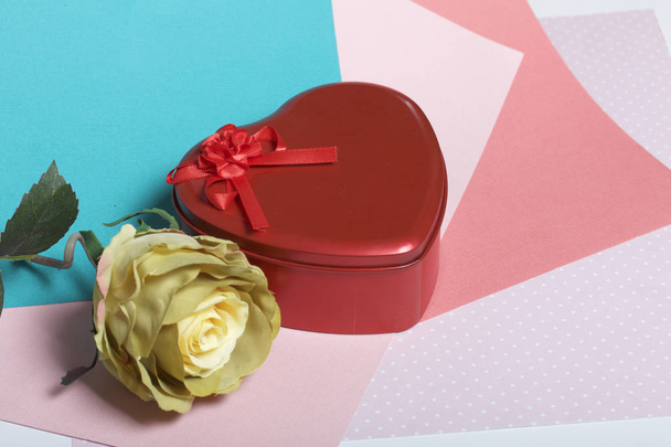 A box in the form of a red heart with a gift. Next is a flower. On a background of multicolored paper. - Foto, Imagem