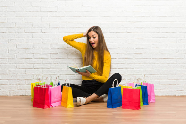 Νεαρό κορίτσι με πολλή έκπληξη απολαμβάνοντας διαβάζοντας ένα βιβλίο τσάντες για ψώνια - Φωτογραφία, εικόνα