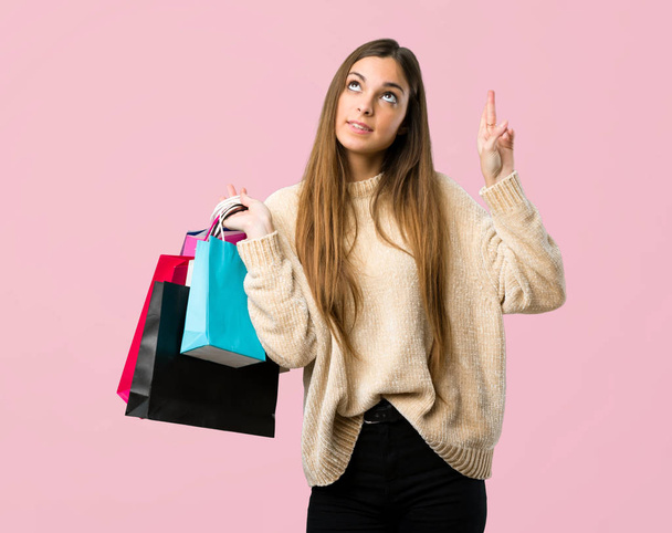 Young girl with shopping bags with fingers crossing and wishing the best on isolated pink background - Photo, Image