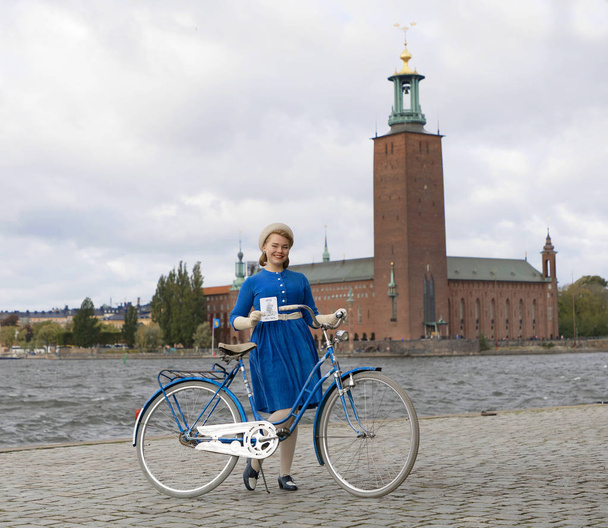 STOCKHOLM - SEPT 22, 2018: Beautiful smiling woman wearing old fashioned blue dress holding a retro bicycle in front of Stockholm City Hall in the Bike in Tweed event September 22, 2018 in Stockholm, Sweden - Photo, image