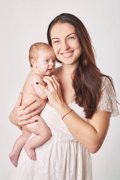 молодая мама с натуральным макияжем держит ребенка на руках, снимая на светлом фоне
 - Фото, изображение