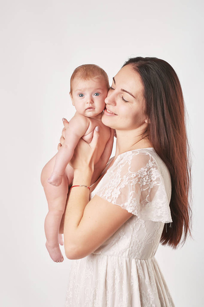 молодая мама с натуральным макияжем держит ребенка на руках, снимая на светлом фоне
 - Фото, изображение