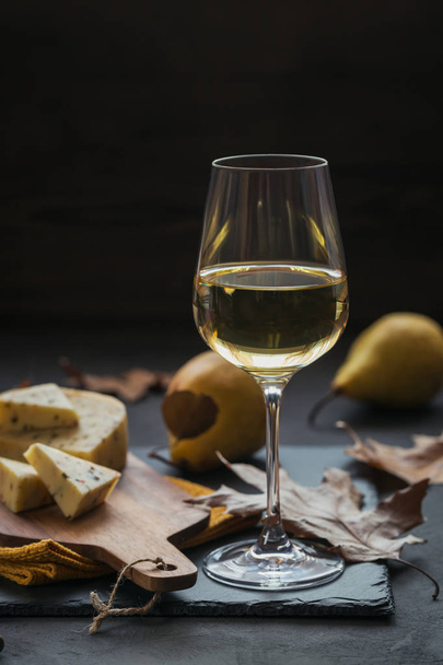 A glass of white wine was served with cheese in a cutting board on dark background. Autumn picnic with cheese, wine and dry leaves in rustic style. - Фото, зображення