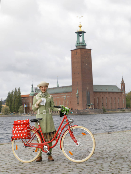 Sztokholm - 22 września 2018: Młoda uśmiechnięta kobieta w staromodnej zielonej sukience z zabytkowym rowerem przed ratuszem w Sztokholmie na rowerze w Tweed 22 września 2018 w Sztokholmie, Szwecja - Zdjęcie, obraz