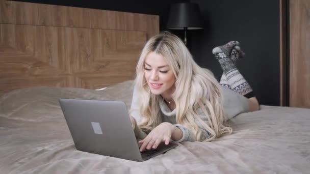 Beautiful blonde woman lying on the bed with gray aluminum laptop. Girl smiles, good mood. Blogging, browsing internet, chatting. In a warm cozy sweater and wool socks. Happy winter concept. - Filmmaterial, Video