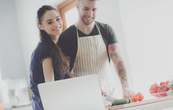 Young man cutting vegetables and woman standing with laptop in the kitchen - Foto, Bild