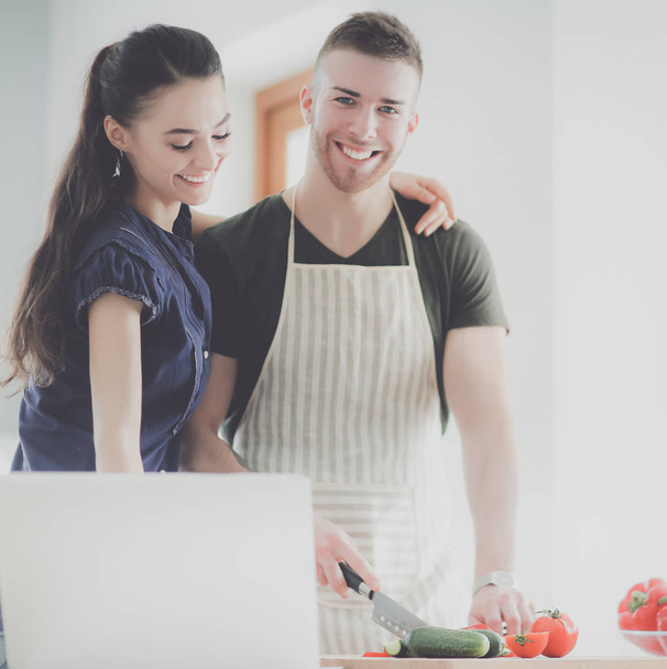 Young man cutting vegetables and woman standing with laptop in the kitchen - Foto, Imagen
