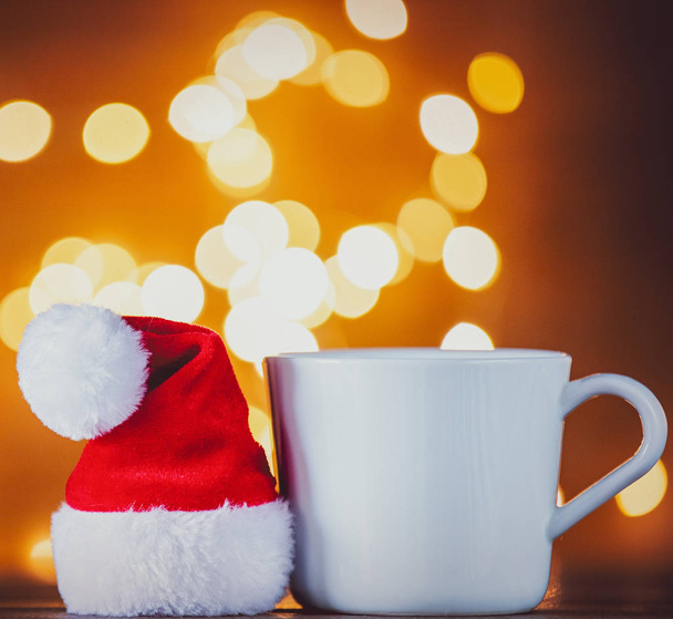 White cup of tea or coffee and Santa Claus hat on Christmas Lights on background - Photo, Image